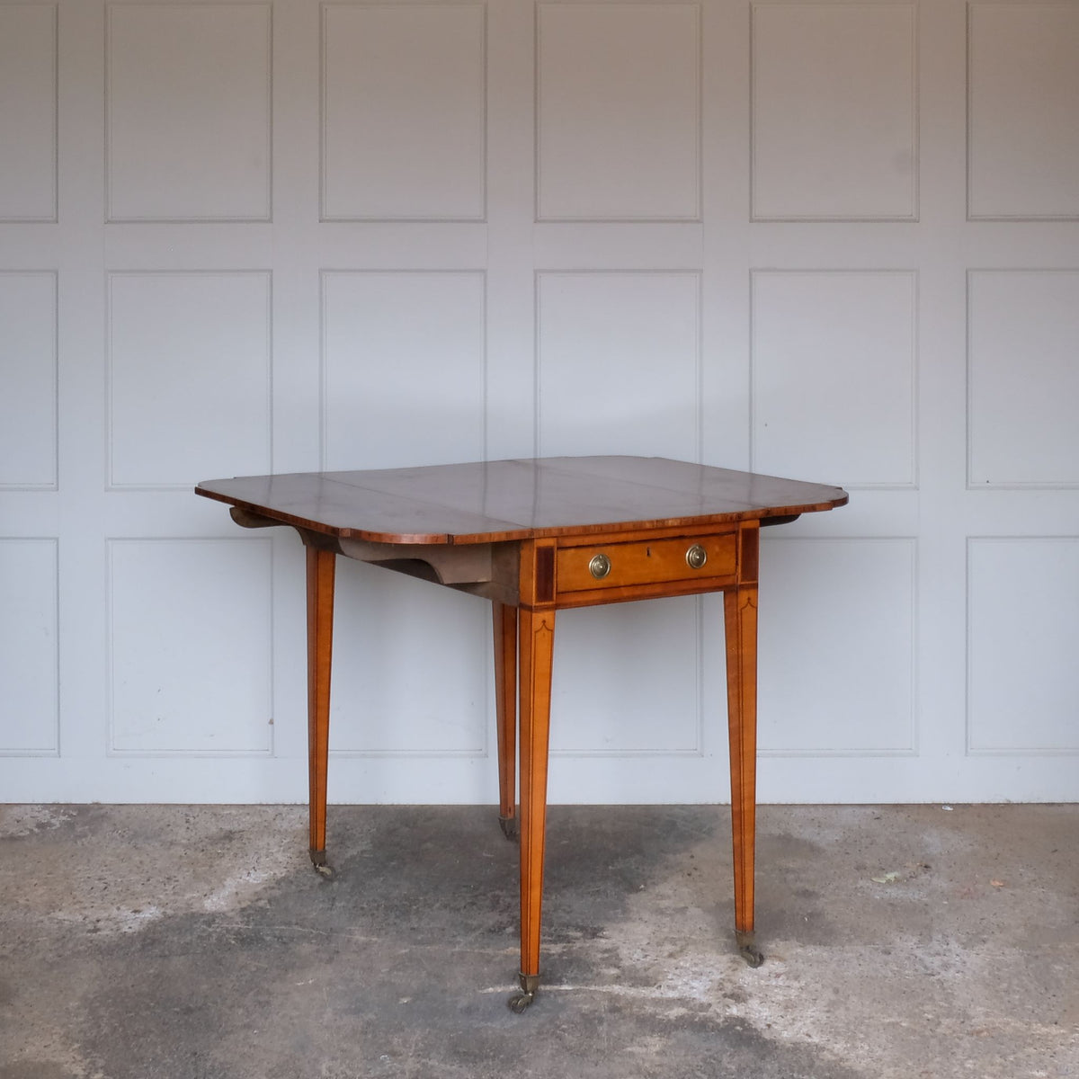 A lovely George III satinwood Pembroke table, the shaped drop leaf top with elegant crossbanding above a single end drawer with a dummy drawer on the opposing side, raised on gently tapering square legs with delicate inlay detailing over brass castors, with some gentle surface patina to the top as is commensurate with age and use, in very good condition
